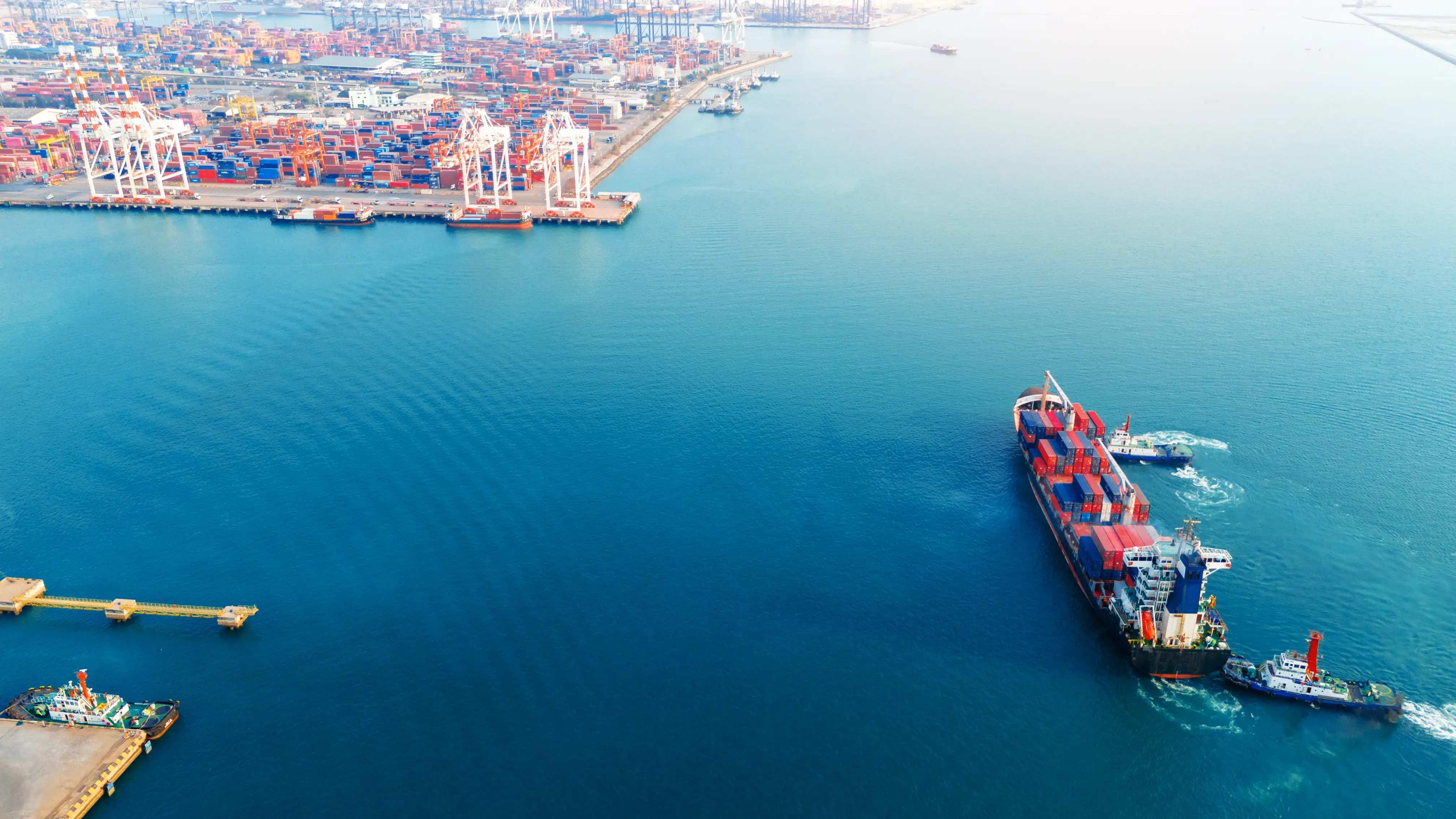 Aerial view of a container ship guided by tugboats toward a port terminal